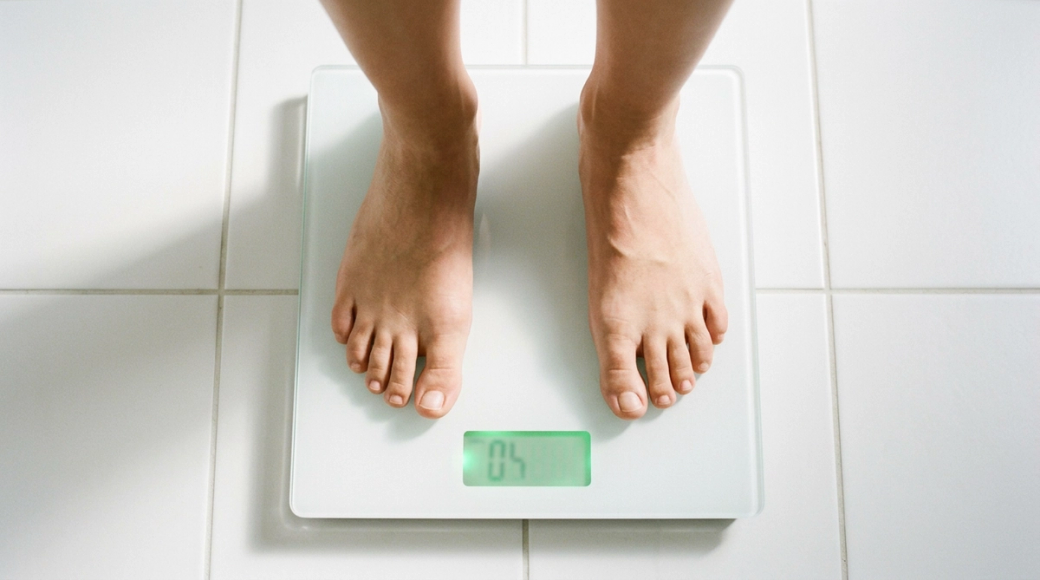 The image showcases a pair of bare feet standing on a digital bathroom scale, viewed from above. The scale's display features a glowing green number that is not legible, set against a clean white tiled floor illuminated by soft morning light, creating a serene and optimistic atmosphere.