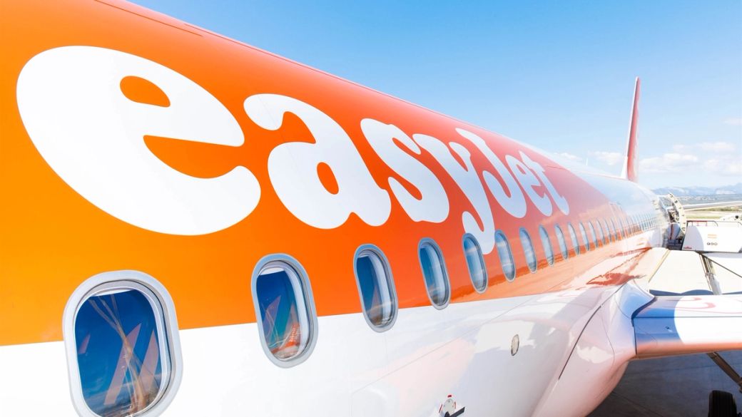 The image features an easyJet airplane with a prominent orange and white fuselage against a clear blue sky. The aircraft is parked on the tarmac, showcasing its branding.