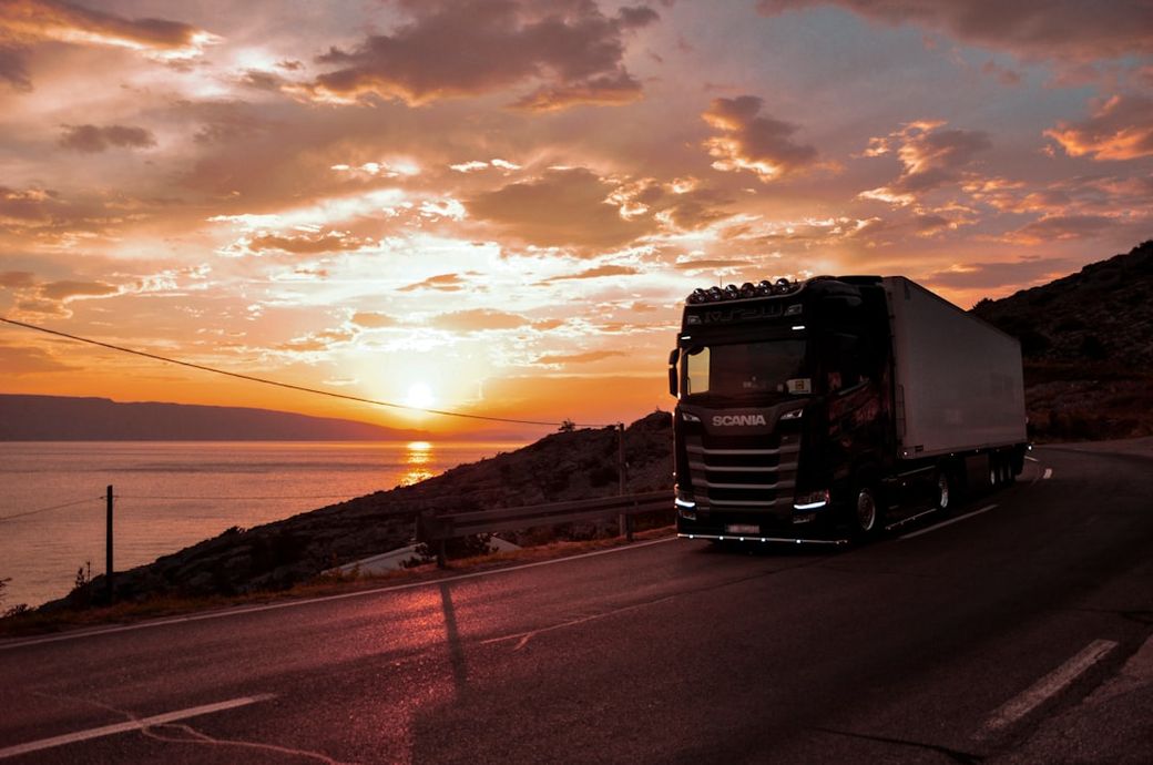 A Scania truck is driving along a coastal road during sunset. The sky is vibrant with orange and pink hues, reflecting over the calm sea, creating a picturesque landscape. — Credit: Photo by Natalia Marcelewicz on Unsplash