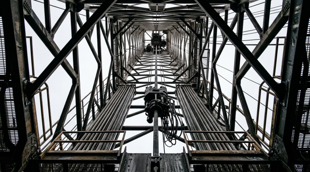 The image captures the interior view of a drilling derrick tower, looking directly upward. The structure's intricate steel lattice forms a geometric pattern converging toward the crown block, silhouetted against a pale overcast sky, while drilling equipment and pipe racks embellish the sides, revealing an industrial environment characterized by heavy machinery.