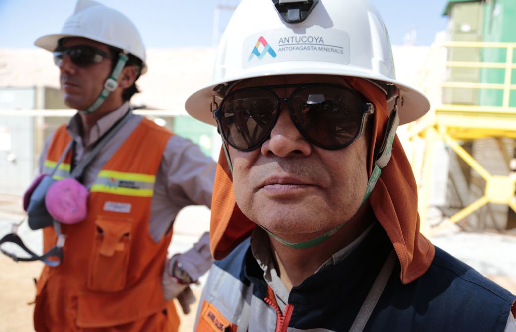 The image features two workers at a mining site. One worker is prominently in focus, wearing a helmet and sunglasses, while another worker is slightly blurred in the background. Both are dressed in safety gear, indicating they are on-site at a mining operation.
