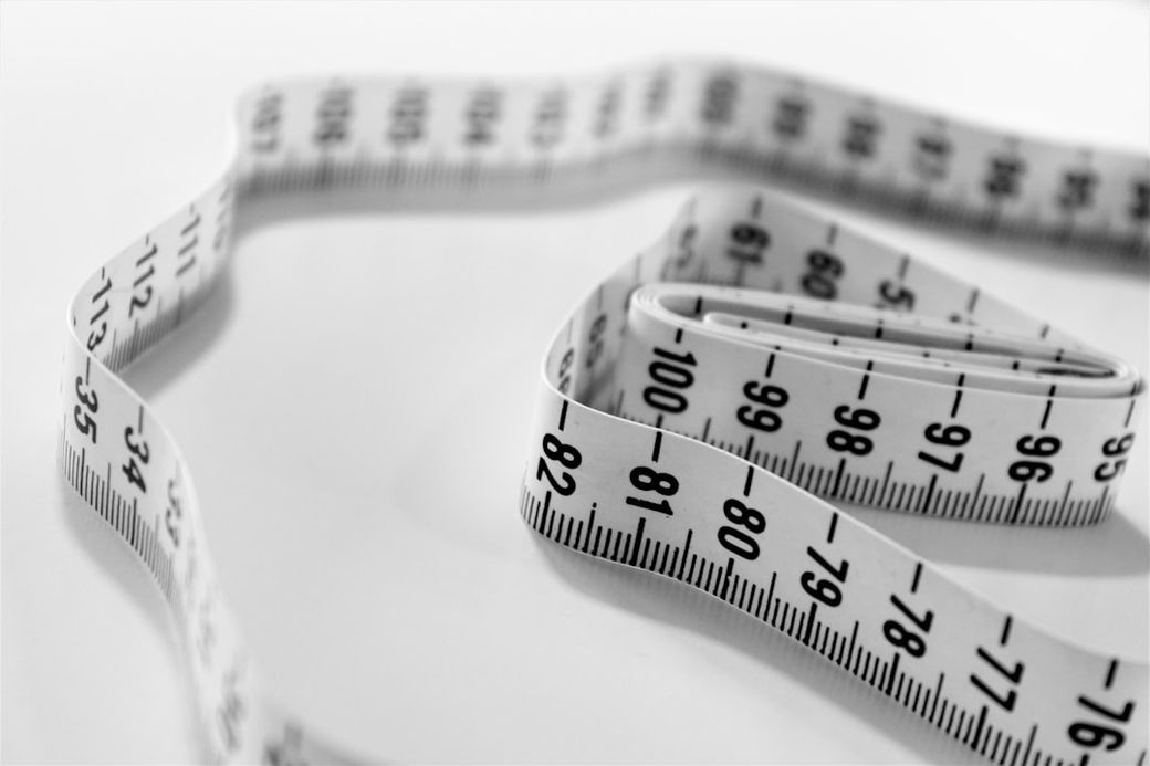 The image shows a flexible measuring tape coiled in a spiral pattern on a white surface. The tape features clearly marked measurements in centimeters and inches. — Credit: Photo by Siora Photography on Unsplash