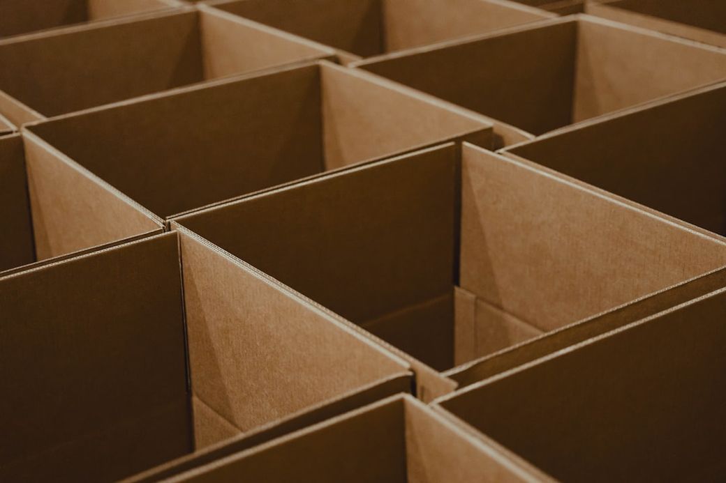 The image shows multiple empty cardboard boxes arranged in a grid pattern. The boxes are uniformly brown and feature open tops, suggesting they are ready for packing or storage. — Credit: Photo by Luke Heibert on Unsplash