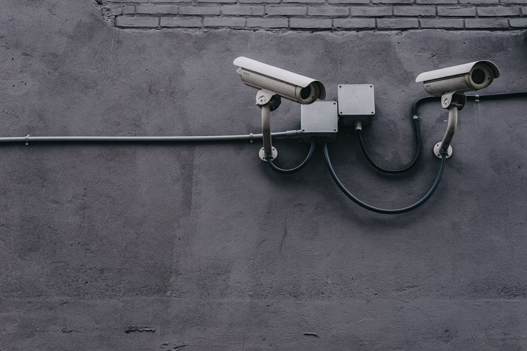 The image features two surveillance cameras mounted on a textured gray wall. The cameras are connected by black cables, emphasizing a focus on security and monitoring. — Credit: Photo by Scott Webb on Unsplash