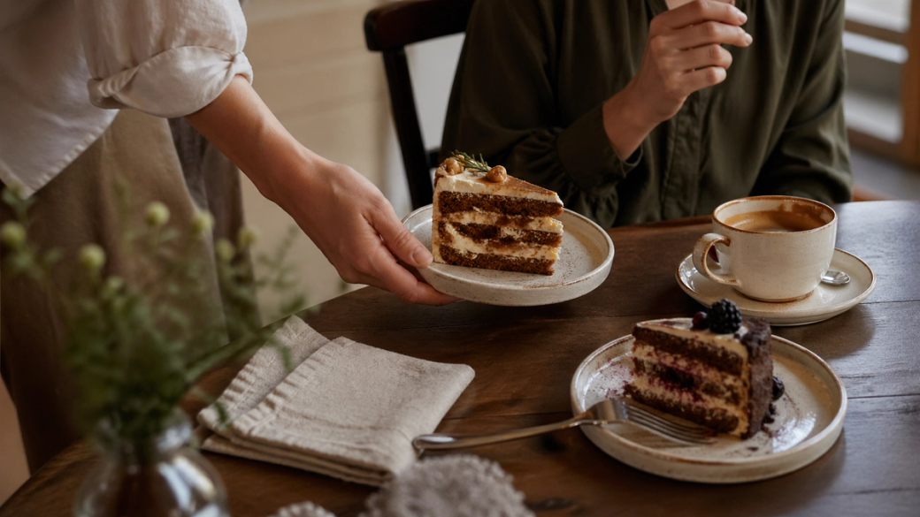 The image depicts a person serving a slice of cake at a cozy café table. Another individual is seated at the table, enjoying a cup of coffee, with two plates of different cake slices displayed on the table.