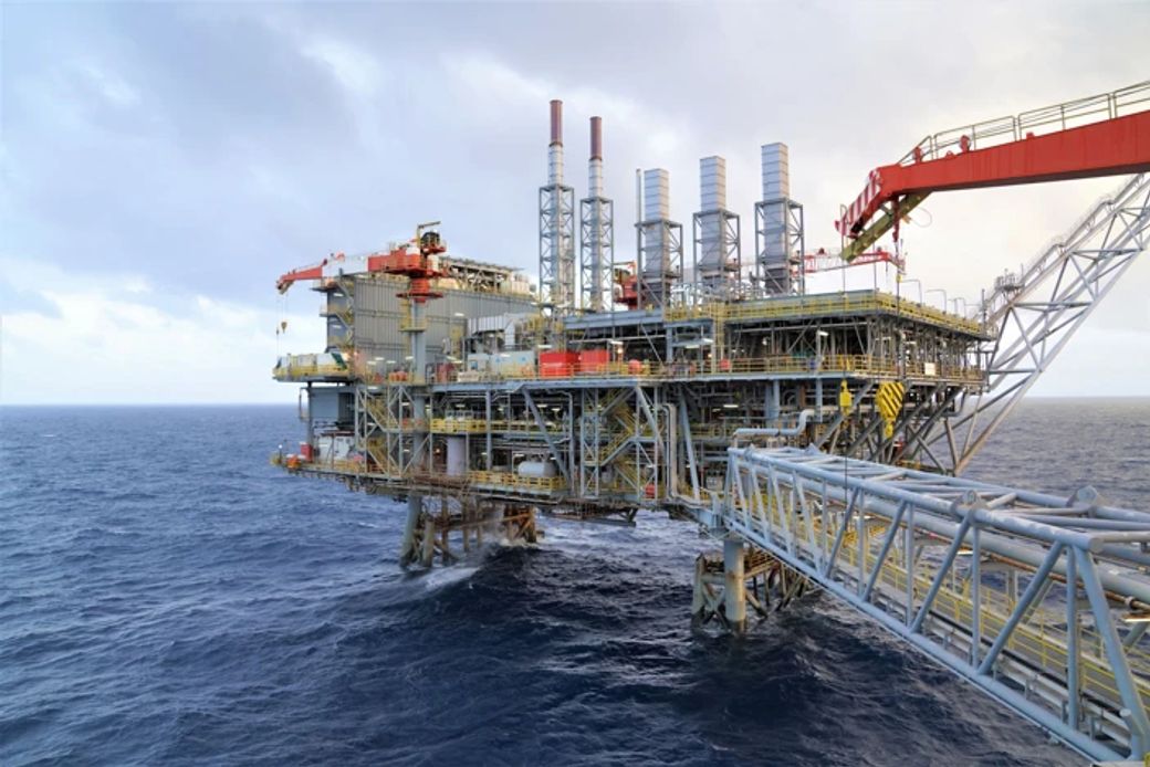 The image shows a large offshore oil rig situated in the ocean. It features multiple structures and equipment, including tall pipes and platforms, indicating an active oil extraction process.