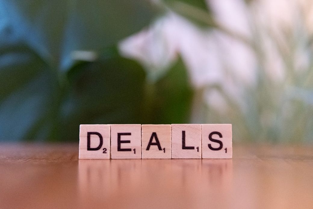 The image features Scrabble-style letter tiles arranged to spell the word 'DEALS'. The background consists of soft green foliage, creating a natural and inviting atmosphere. — Credit: Photo by Markus Winkler on Unsplash