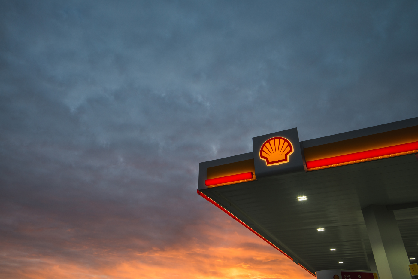 A Shell gas station is prominently displayed under a dramatic sky at sunset. The station's logo and lighting create a striking visual against the colorful clouds.