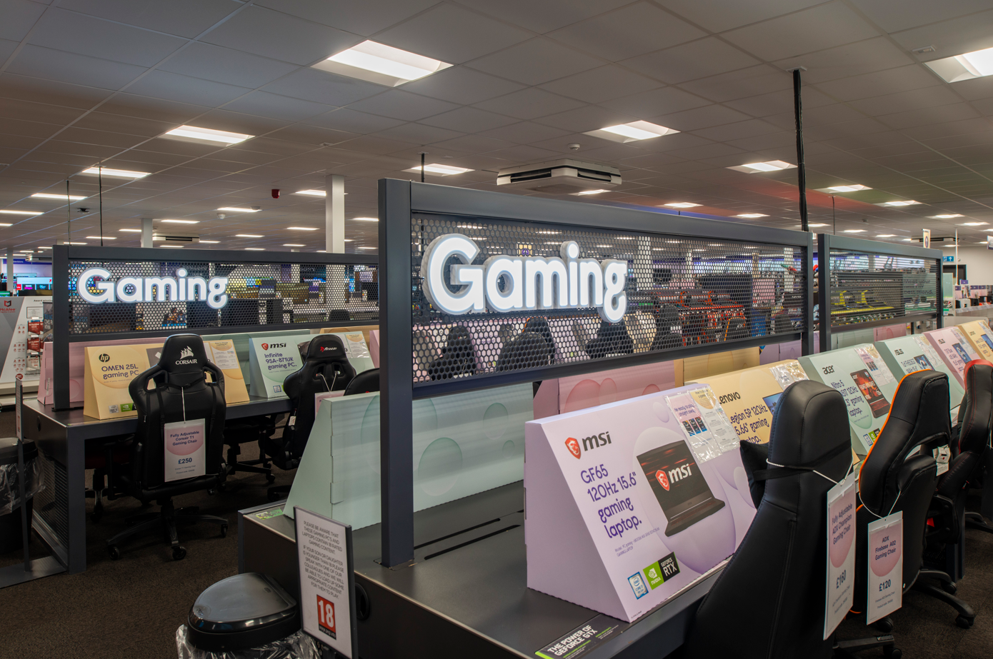 An interior view of a gaming section in a retail store, featuring multiple gaming chairs and signage highlighting 'Gaming.' Several promotional posters for gaming laptops are visible on the displays.