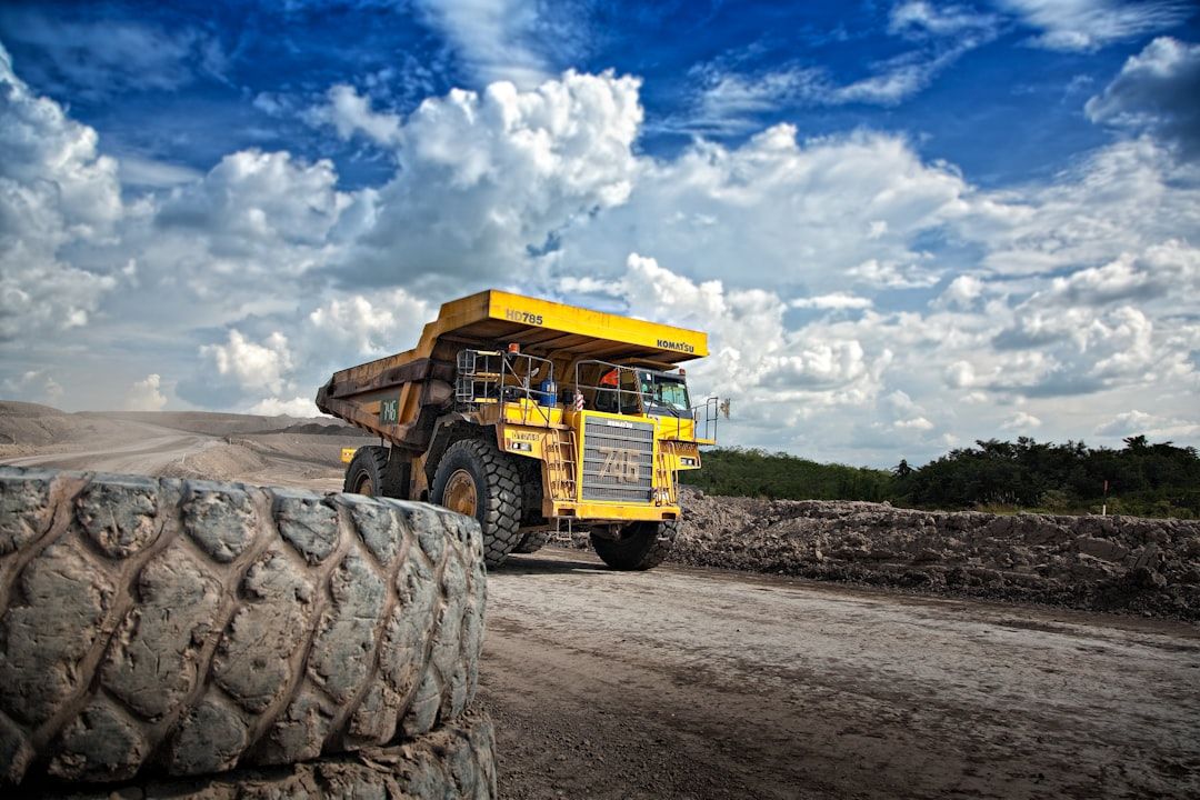 A large yellow dump truck is navigating through a construction or mining site. The sky is filled with clouds, and the terrain appears to be dusty with earth being moved around.