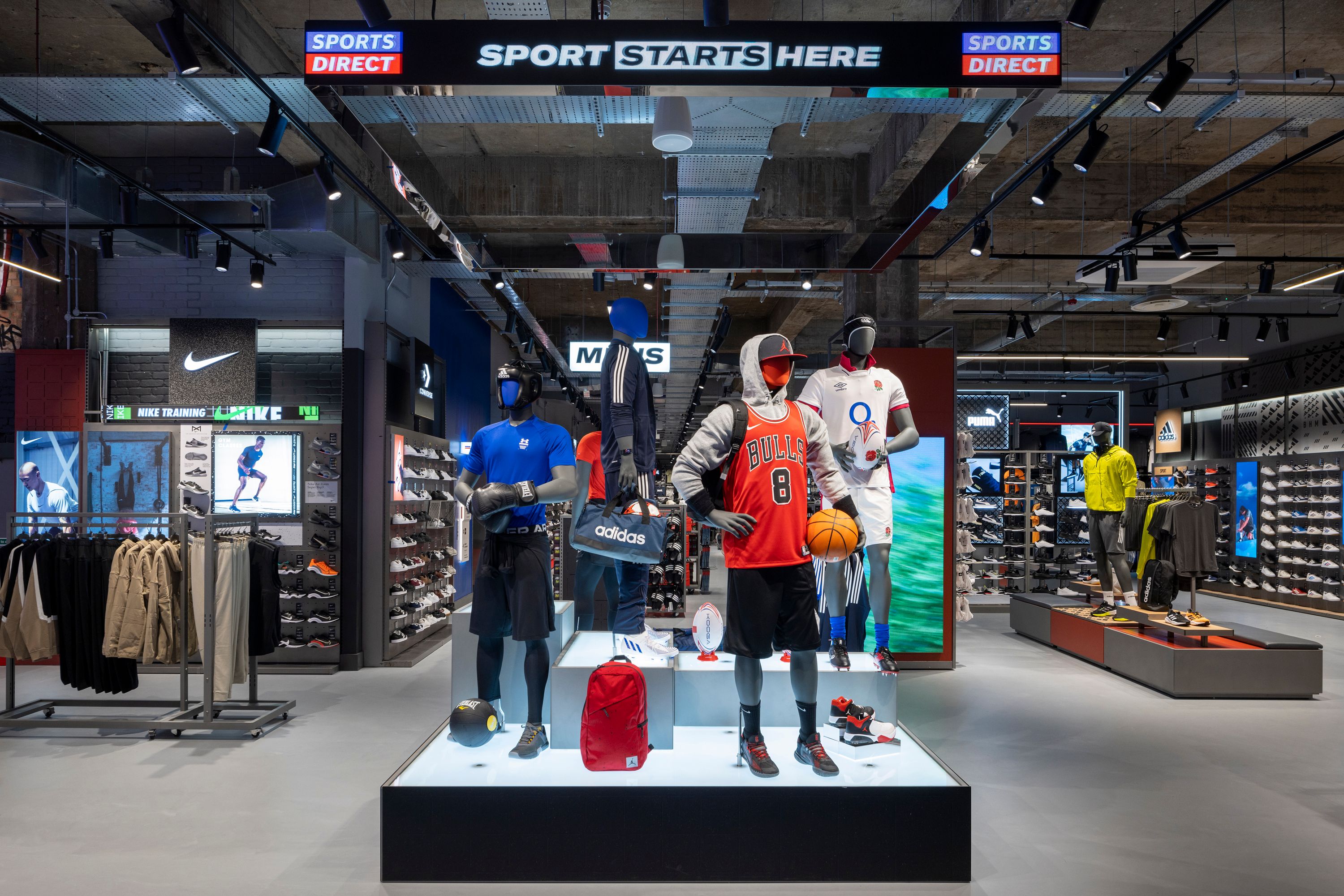 The image showcases a modern sports retail store interior featuring various athletic apparel and footwear. Mannequins are displayed in sports gear, highlighting brands like Adidas, with an emphasis on basketball and athletic performance.