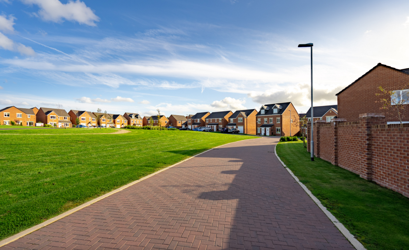 The image shows a residential street featuring several modern houses arranged along a curved path. The scene is set in a suburban area with well-maintained lawns and clear skies.