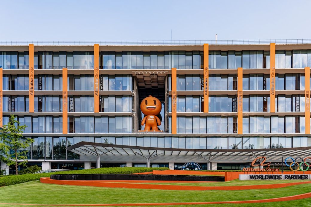 The image shows the headquarters of Alibaba Group featuring a large orange mascot figure prominently displayed in front of the building. The architecture is modern with a green landscape in the foreground. — Credit: Photo by Zonghe Ma on Unsplash