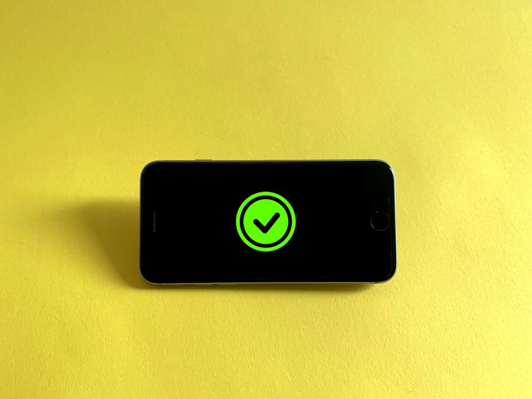 The image depicts a smartphone laying flat on a yellow background. The screen displays a large green check mark, suggesting a confirmation or successful status. — Credit: Photo by Franck on Unsplash