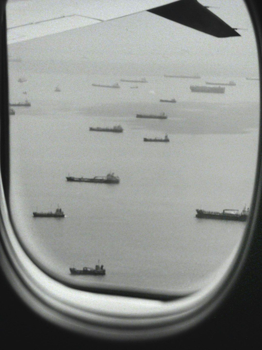 This image captures a view from an airplane window, showcasing a maritime scene with several cargo ships scattered across the water. The monochromatic tone adds a vintage feel to the composition.