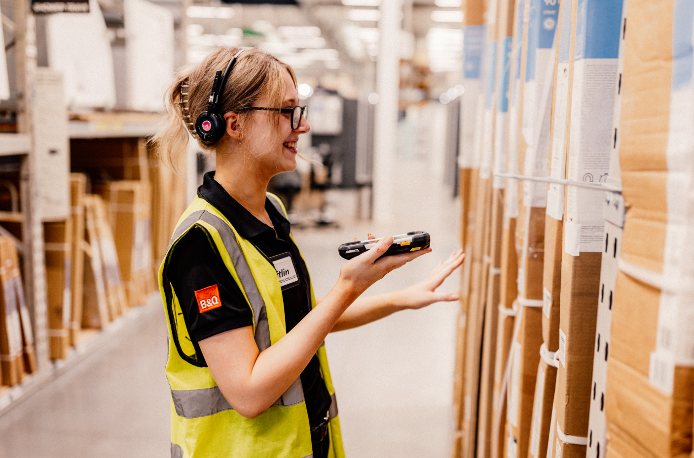 A B&Q retail employee in a high-visibility vest interacts with inventory in a warehouse or home improvement store. She wears a headset and is using a handheld device to scan items, exemplifying modern retail operations.