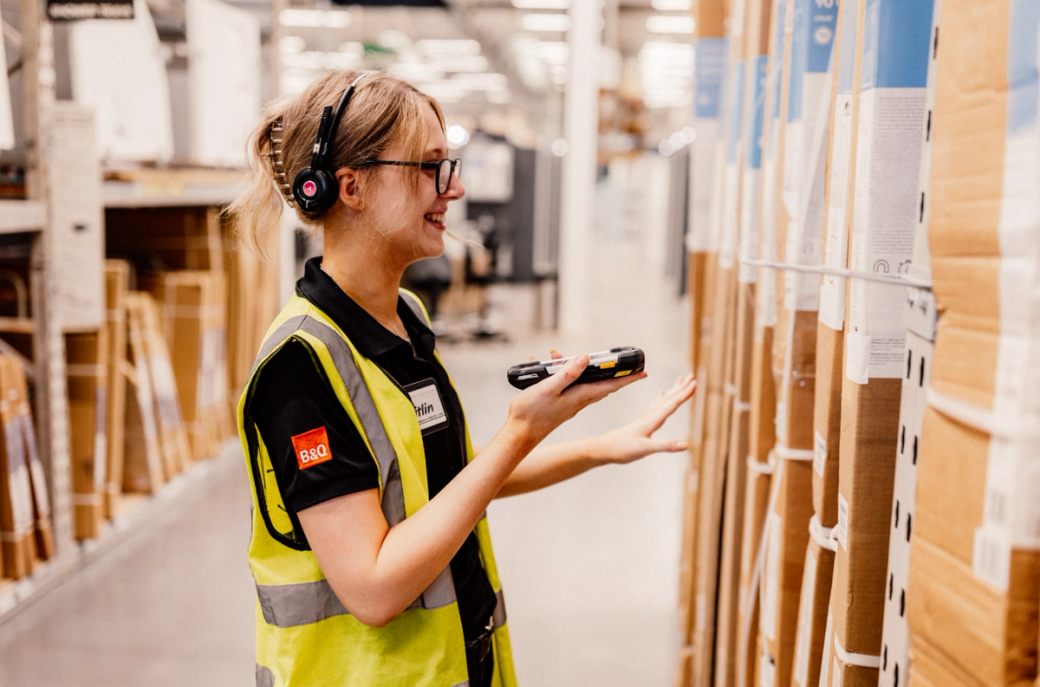 A B&Q retail employee in a high-visibility vest interacts with inventory in a warehouse or home improvement store. She wears a headset and is using a handheld device to scan items, exemplifying modern retail operations.