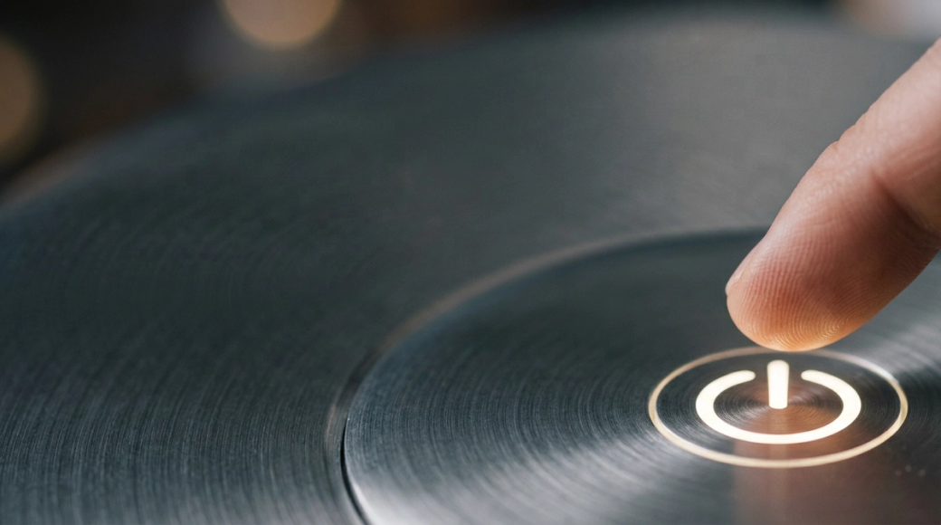 The image captures an abstract macro shot of a circular power button symbol backlit in soft white against a brushed aluminum surface. A fingertip hovers just above the symbol, illustrating the moment before activation, with a shallow depth of field highlighting the glowing symbol and the fine details of the metal.
