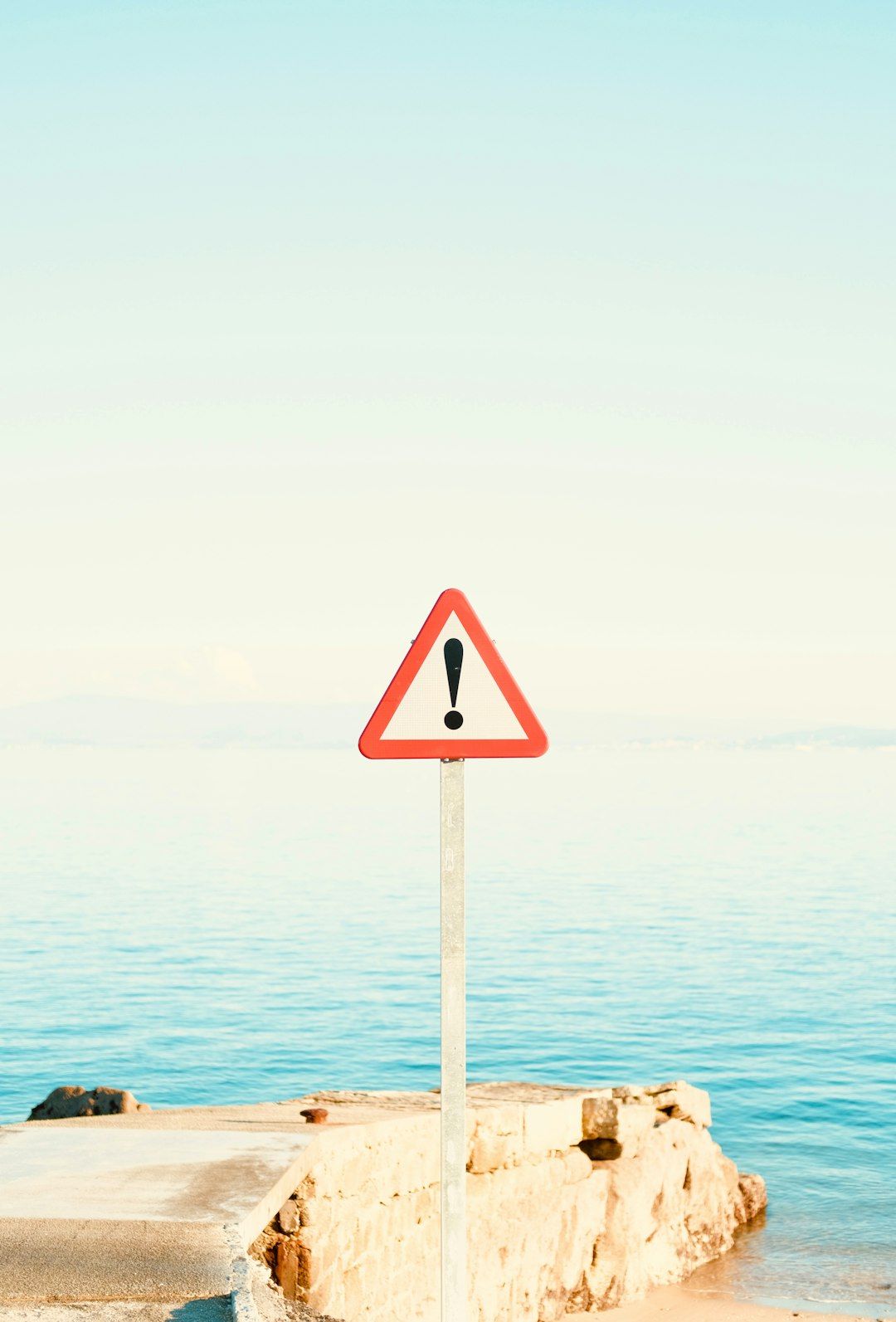 The image depicts a red triangular warning sign with an exclamation mark positioned on a metal pole near a serene body of water. The horizon features a clear blue sky with a slight haze, indicating a tranquil atmosphere.