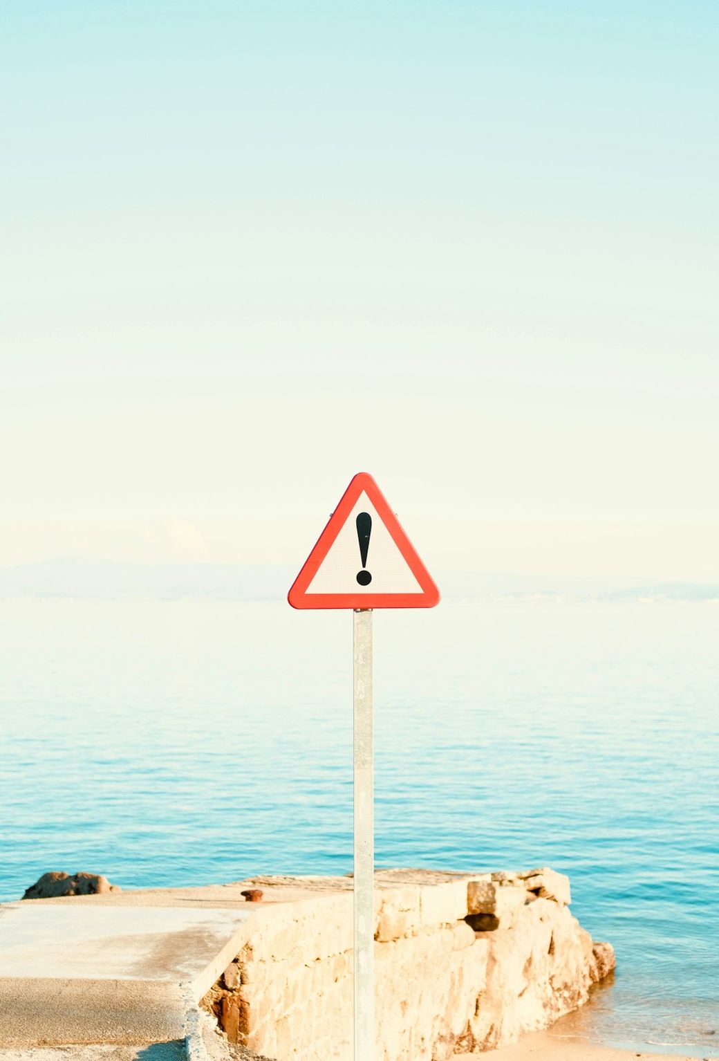 The image depicts a red triangular warning sign with an exclamation mark positioned on a metal pole near a serene body of water. The horizon features a clear blue sky with a slight haze, indicating a tranquil atmosphere. — Credit: Photo by Alex varela on Unsplash