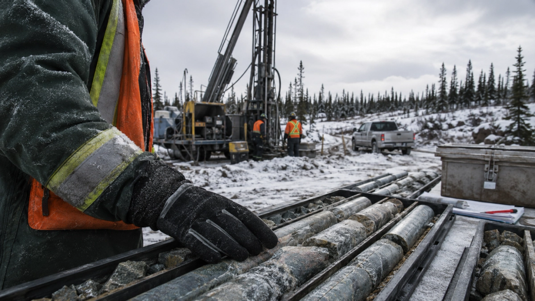 A worker in winter attire inspects metal drill rods on a snowy site where drilling equipment is visible. Two other individuals in the background are operating machinery amidst a forested area.