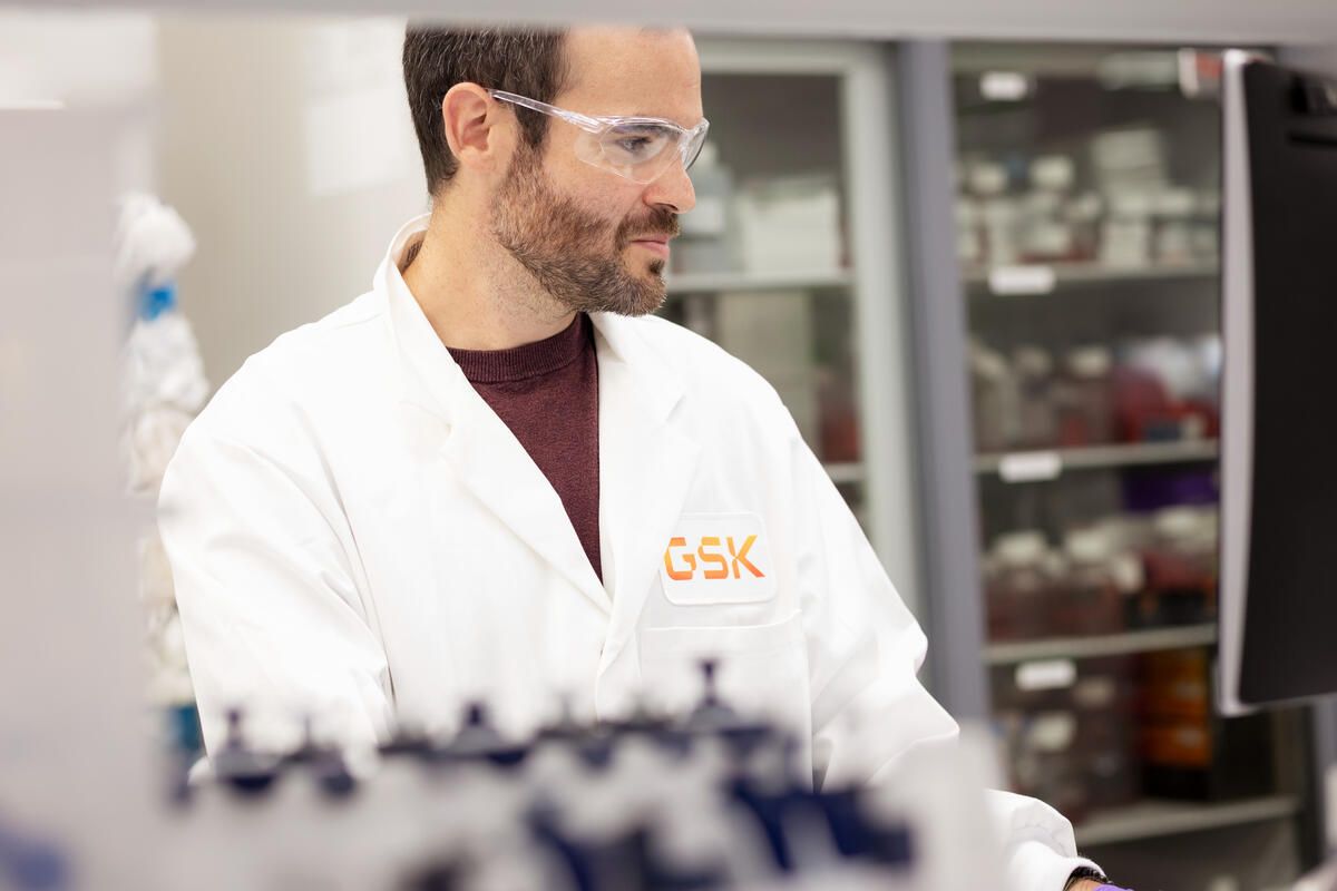 A laboratory technician in a white lab coat is working at a computer in a laboratory setting. The technician is wearing safety goggles and appears focused on the task at hand, with laboratory shelves filled with various samples in the background.