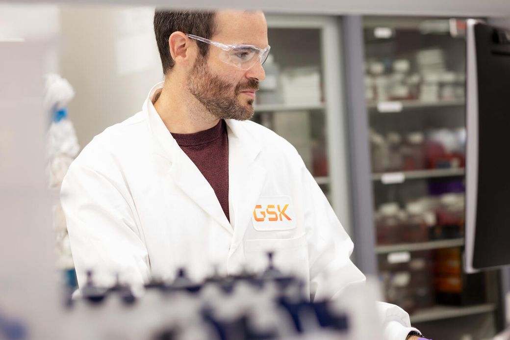 A laboratory technician in a white lab coat is working at a computer in a laboratory setting. The technician is wearing safety goggles and appears focused on the task at hand, with laboratory shelves filled with various samples in the background.