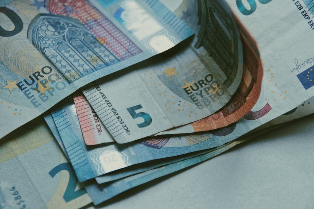 The image features a close-up view of various Euro banknotes, including denominations of 5, 20, and 50 euros, stacked together on a neutral background. The notes display intricate designs typical of Euro currency, showcasing their colors and features.