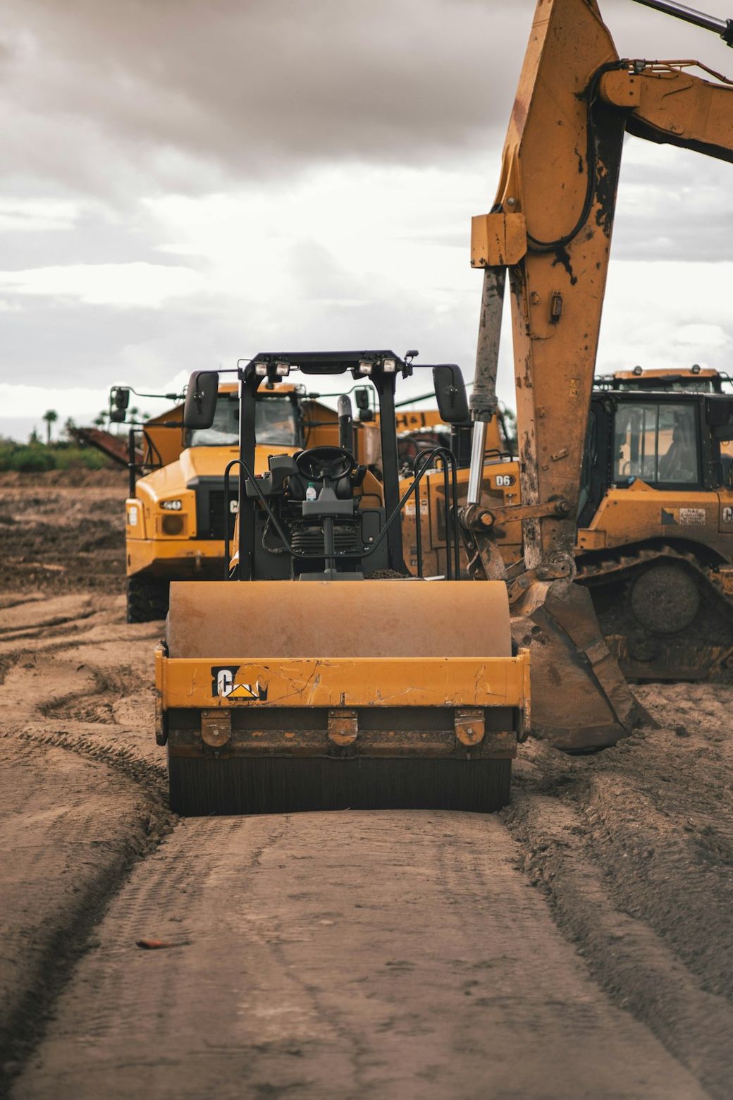 The image depicts heavy construction machinery including bulldozers and rollers on a dirt road. The cloudy sky suggests an overcast day, and the equipment is arranged in a construction site setting. — Credit: Photo by Jason Jarrach on Unsplash