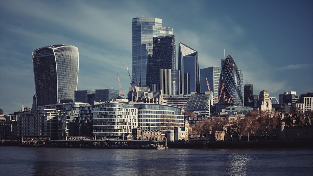 The image showcases a skyline view of London's financial district, featuring a mix of modern skyscrapers and notable architectural designs against a clear blue sky. The scene captures the vibrant urban environment along the River Thames. — Credit: Photo by Tadas Petrokas on Unsplash