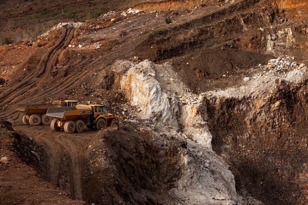 The image depicts a mining site with large machinery at work. Two yellow dump trucks are positioned on a rugged terrain, showcasing the excavation of rocky soil. — Credit: Photo by MiningWatch Portugal on Unsplash