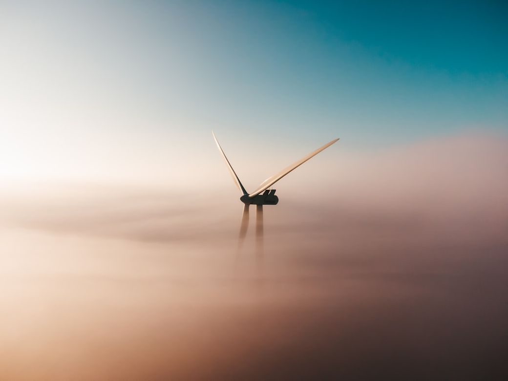 The image portrays a wind turbine emerging from a sea of fog, set against a blue sky. The blades of the turbine are clearly visible and appear to be in motion, capturing the essence of renewable energy. — Credit: Photo by Sander Weeteling on Unsplash