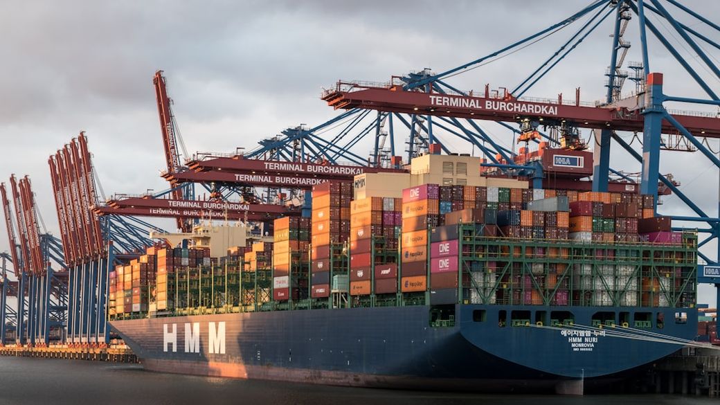 A large container ship named HMM is docked at the Terminal Burchardkai, surrounded by towering crane structures. The ship is loaded with colorful shipping containers, ready for unloading at the busy port. — Credit: Photo by Wolfgang Weiser on Unsplash