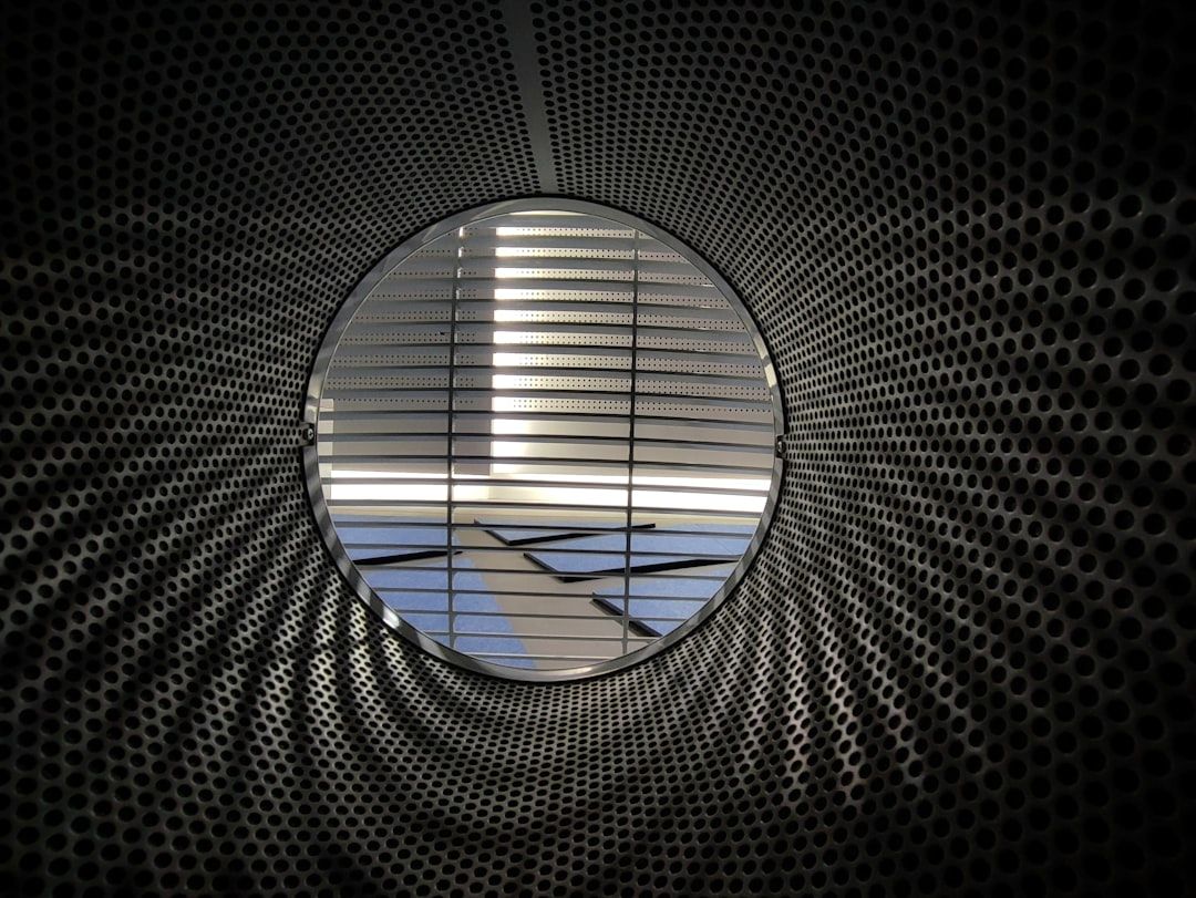 The image features a circular view from the inside of a metallic structure with a perforated pattern. Light filters through the openings, creating a dynamic interplay of shadows and reflections on the interior surface.