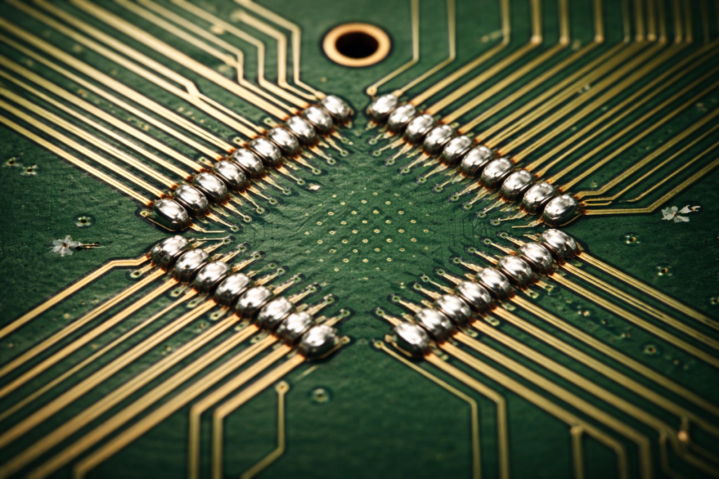 The image showcases a close-up view of a printed circuit board featuring intricate gold wiring and soldered connections. The focus is on the central chip area, highlighting the complexity of modern electronics.