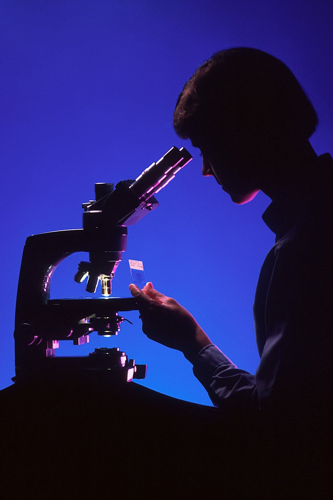 A silhouette of a person examining a sample under a microscope with a glowing background. The image conveys a sense of scientific inquiry and research.