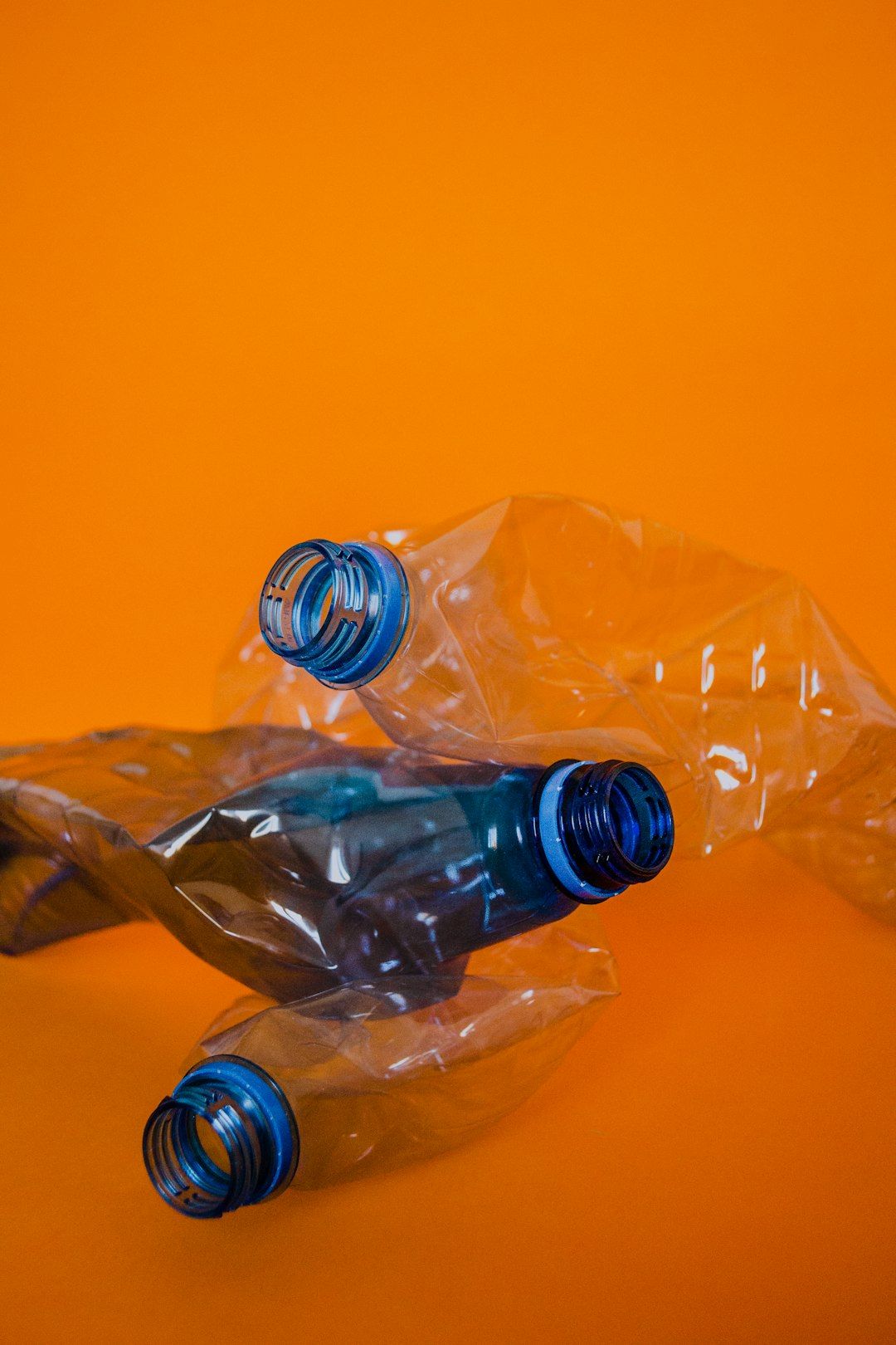 The image features crumpled, transparent plastic waste, including two blue bottle caps, set against a vibrant orange background. This visual highlights environmental issues related to plastic pollution.
