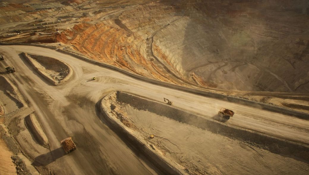 An aerial view of a large open-pit mining site, showcasing the layered earth tones of the excavated terrain. Heavy machinery is visible on the roads within the pit, indicating active mining operations.