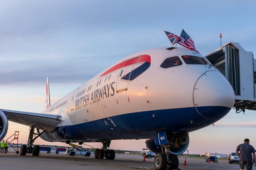 A British Airways aircraft, a Boeing 787, is parked at an airport gate with a sunset sky in the background. The airplane features the British flag on its nose and is being prepared for boarding.