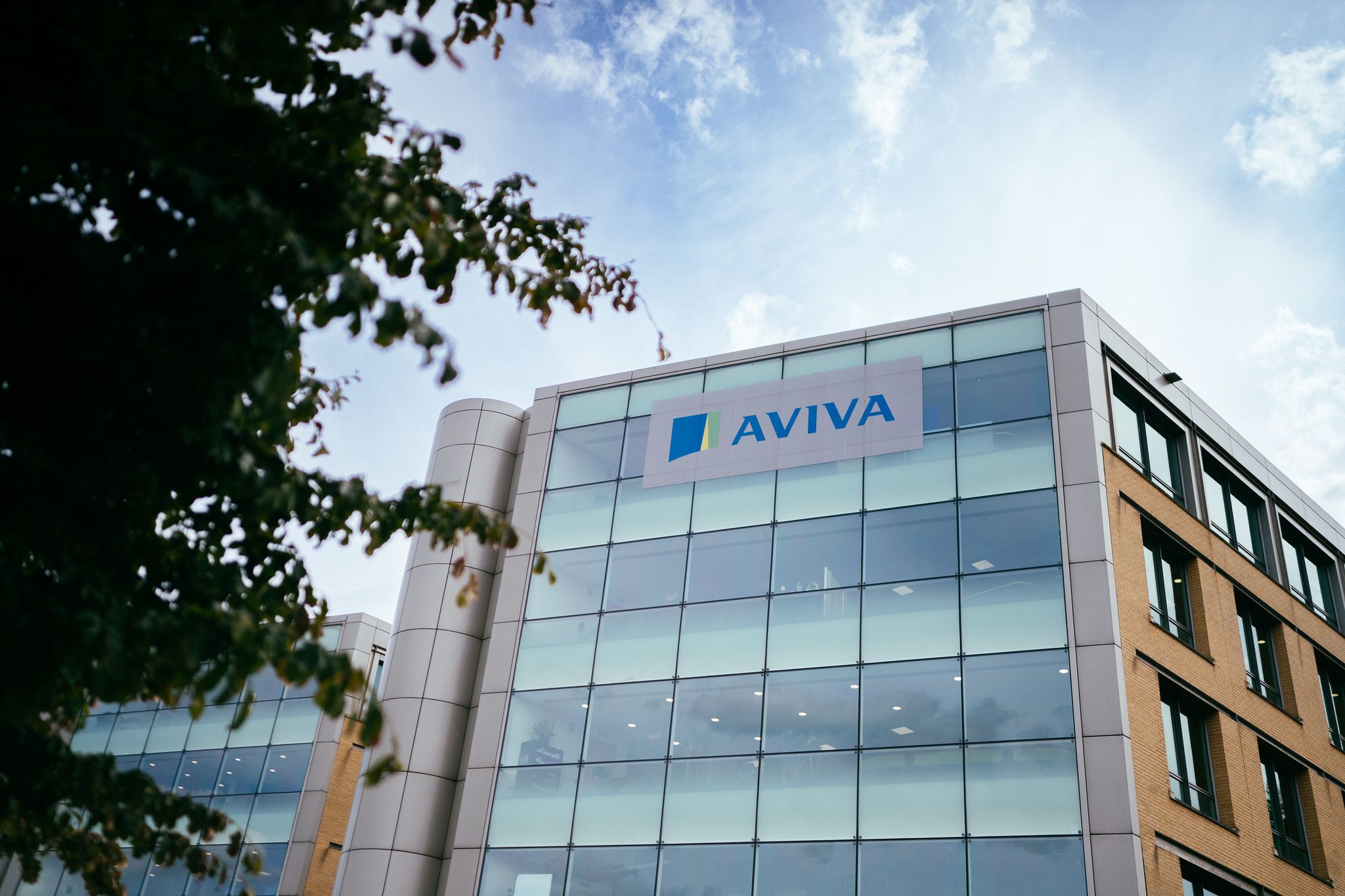 The image shows a modern office building with large glass windows and a visible brand logo on the facade. The logo is from Aviva, a multinational insurance company, and the setting includes a clear blue sky with wispy clouds.
