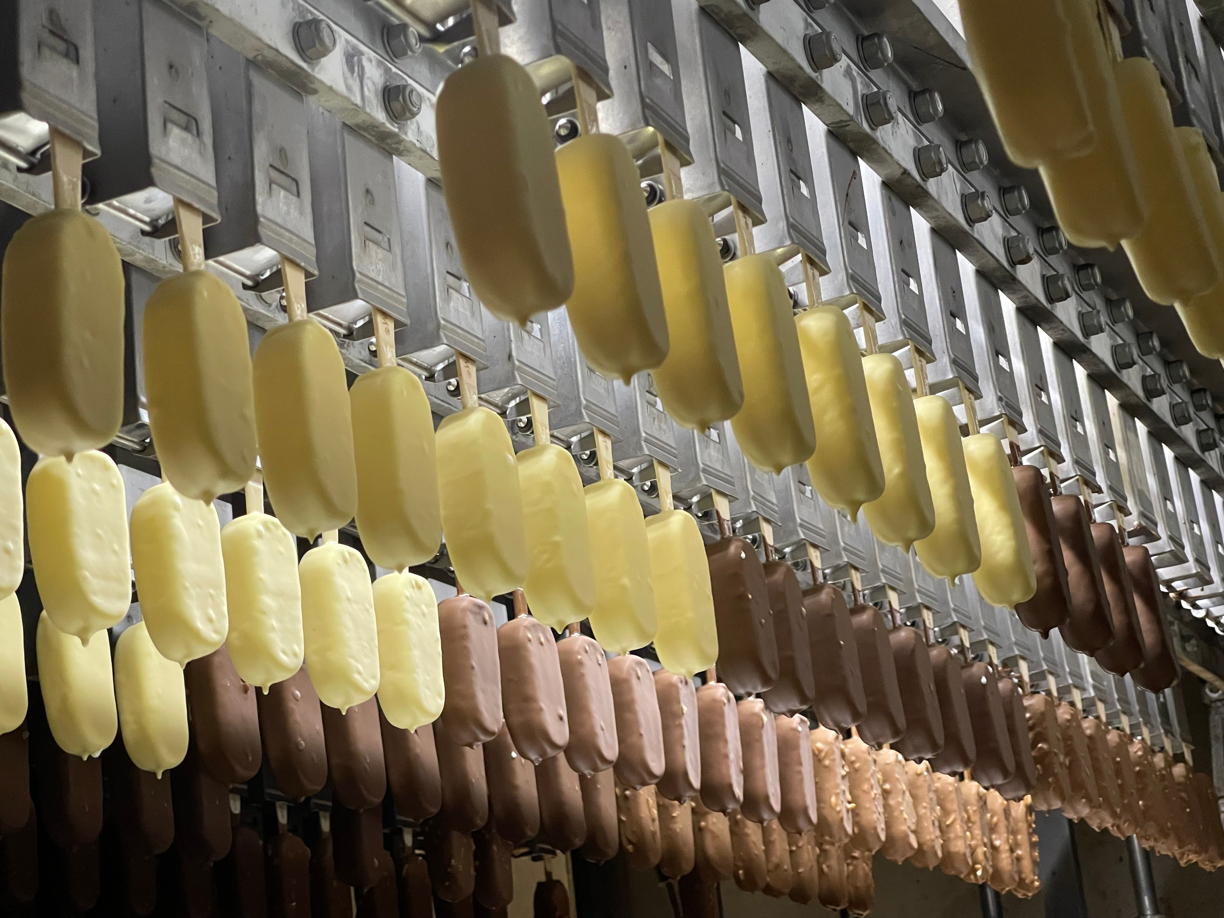 The image depicts a production line of ice cream bars in a factory setting, showcasing various stages of processing. The bars are hanging from a conveyor system, with some coated in yellow chocolate and others in brown chocolate.