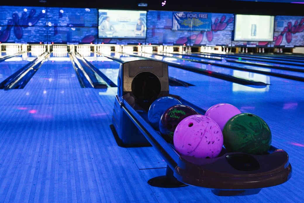 An interior view of a bowling alley featuring multiple bowling lanes and a ball return system. The scene is illuminated with colorful lights and screens displaying bowling graphics.