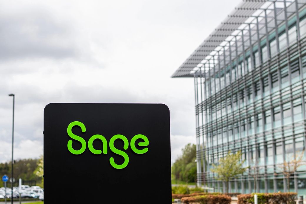 The image shows a modern building with distinctive architecture, featuring a prominent sign displaying the word 'Sage' in green lettering. The setting appears to be an office complex surrounded by greenery and gray clouds overhead.
