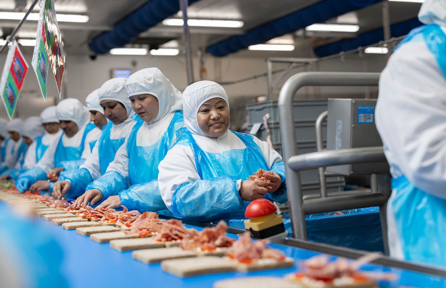 The image shows workers in protective clothing assembling food products in a factory setting. The individuals, predominantly women, are focused on their tasks at a conveyor belt. The environment reflects a food processing or packaging facility.