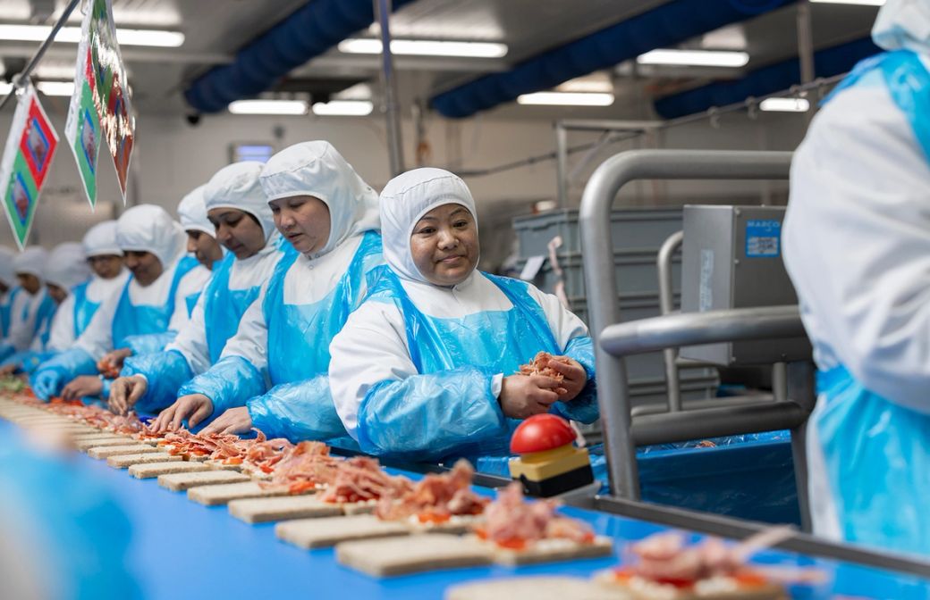 The image shows workers in protective clothing assembling food products in a factory setting. The individuals, predominantly women, are focused on their tasks at a conveyor belt. The environment reflects a food processing or packaging facility.