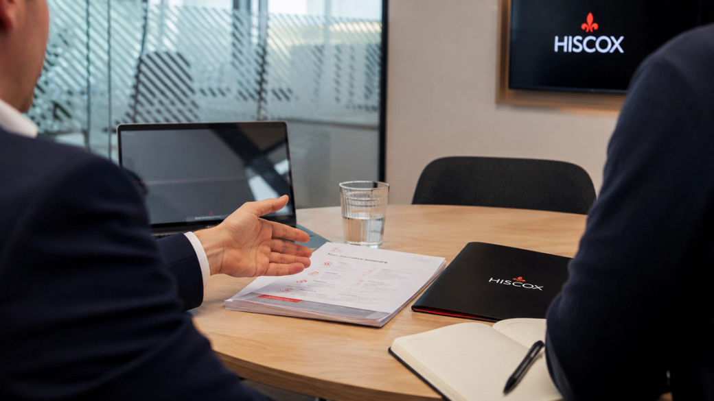 The image depicts a business meeting in a modern office setting, focusing on two individuals discussing documents placed on a conference table. A laptop is open, and a glass of water is visible alongside branded materials from Hiscox.