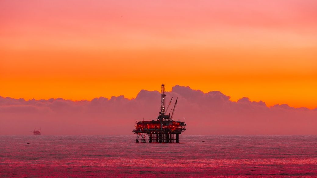 An offshore oil rig is silhouetted against a vibrant sunset sky. The scene captures the tranquil beauty of the ocean at dusk, with the rig's lights glowing against the darkening horizon. — Credit: Photo by Arvind Vallabh on Unsplash