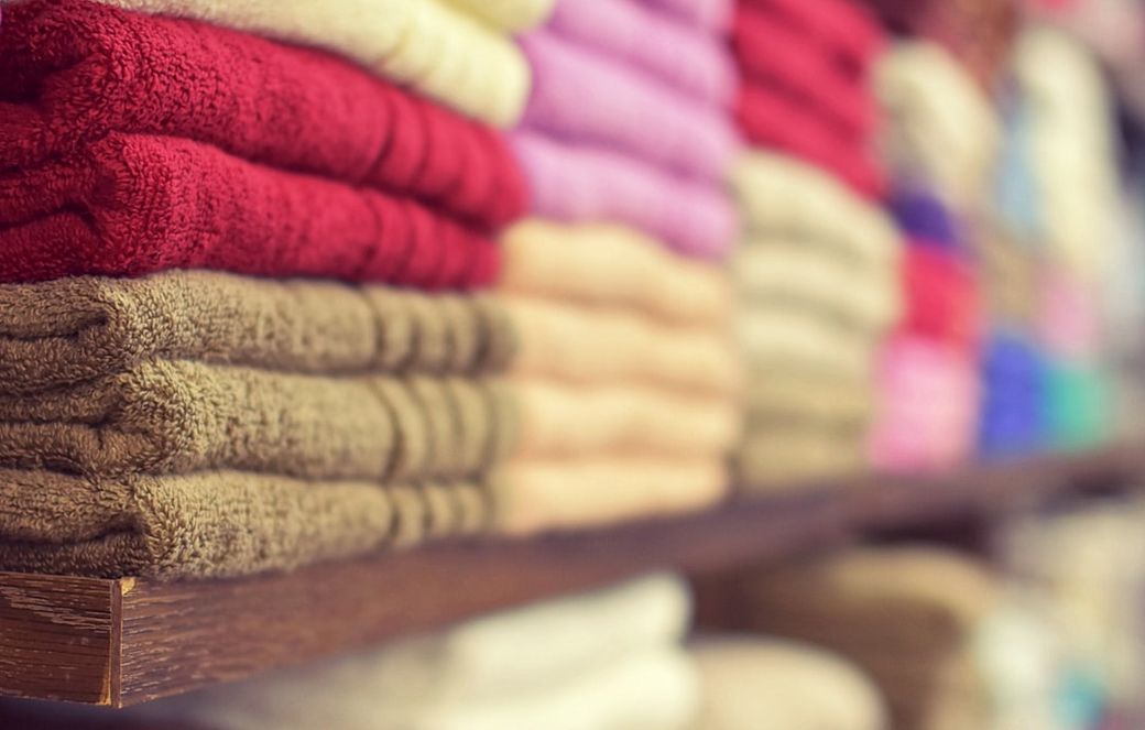 The image features neatly stacked towels in various colors on a wooden shelf. The arrangement creates a visually appealing display of textiles commonly found in retail settings. — Credit: Photo by LumenSoft Technologies on Unsplash