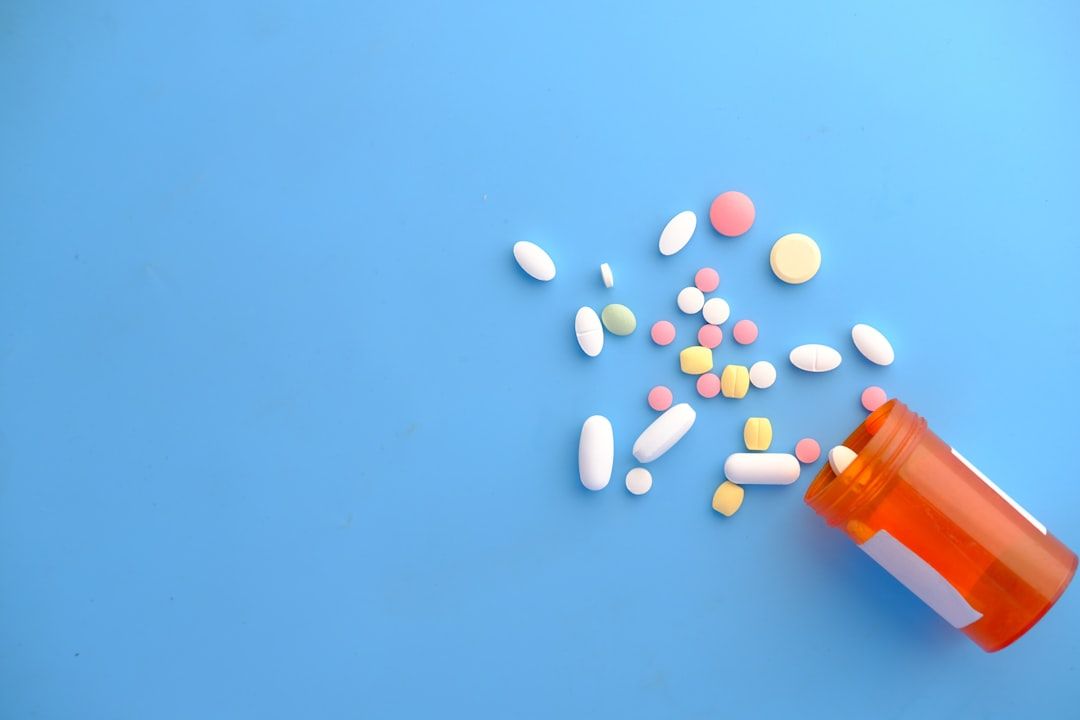 The image shows a variety of prescription and over-the-counter medications scattered around an orange pill bottle. The setting features a blue background, emphasizing the colorful assortment of pills.