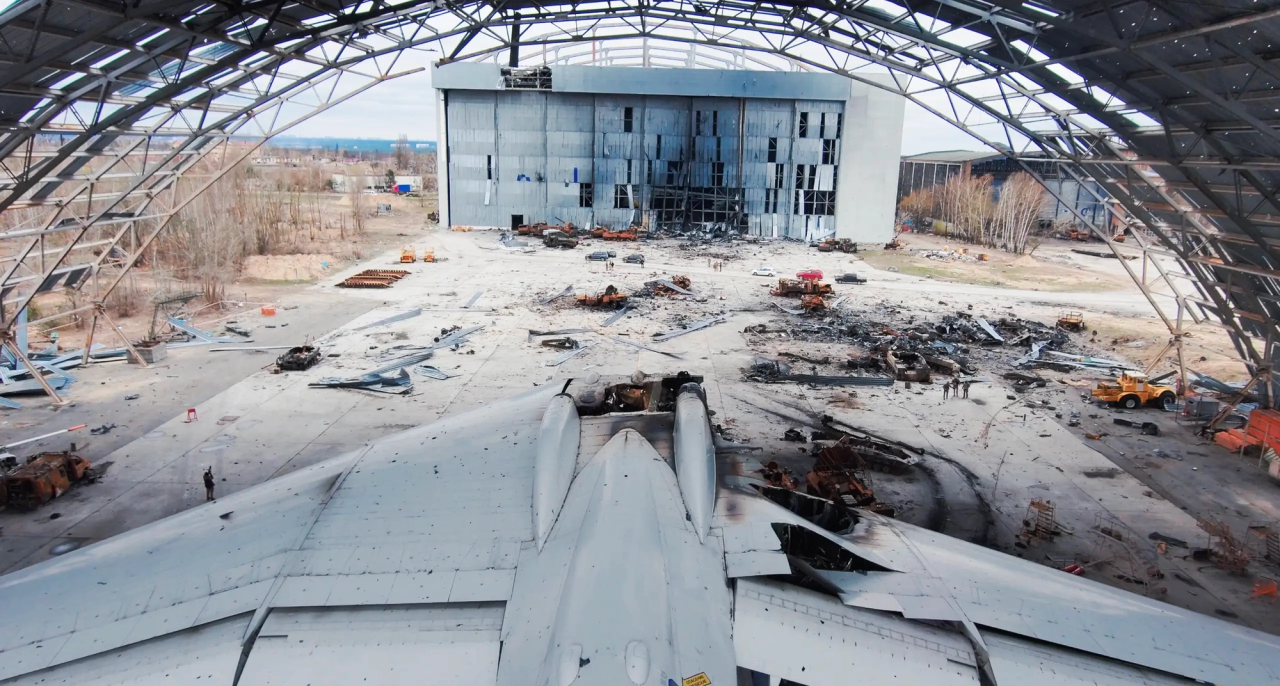 Wide lens photo of an airport hangar, forground at bottom of frame is an aircraft that sustained damage as well as some debris in the background.